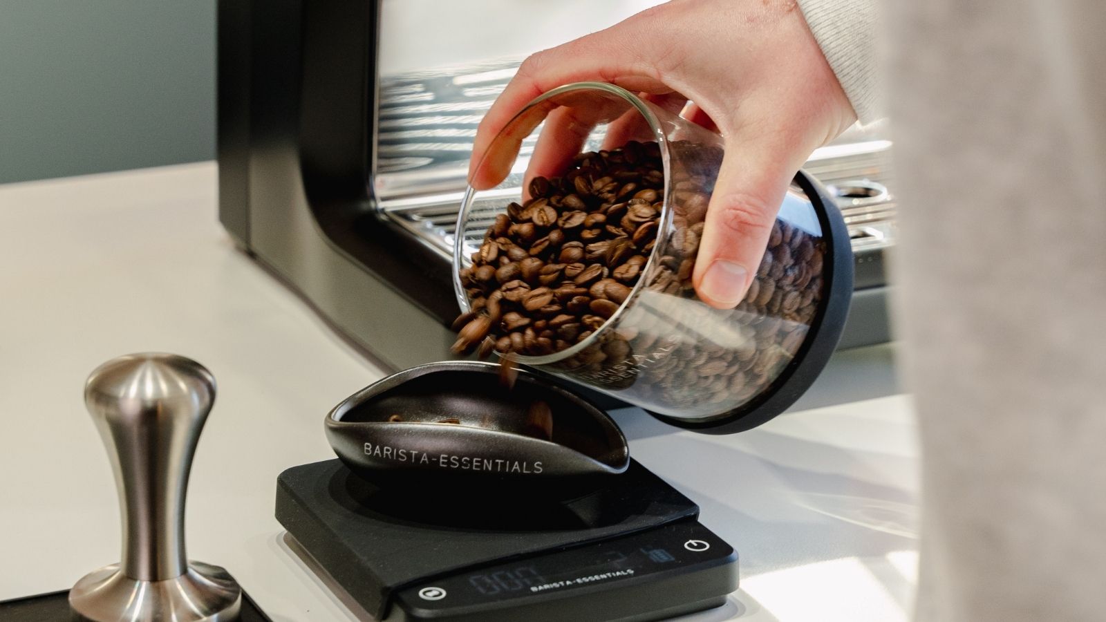 Koffiebonen worden uit een glazen bewaarpot in een doseerbakje op een koffieweegschaal van Barista-Essentials geschonken.