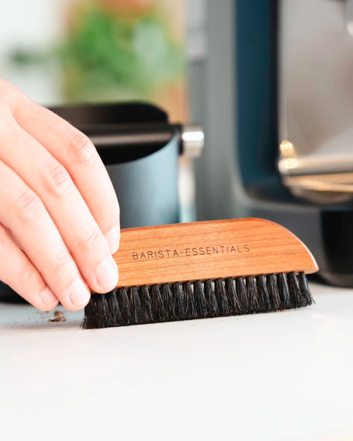 Afbeelding van een hand die een houten borstel met zwarte haren vasthoudt, met de tekst "BARISTA-ESSENTIALS" erop gegraveerd. De borstel wordt gebruikt om koffiedik van een werkblad te vegen.